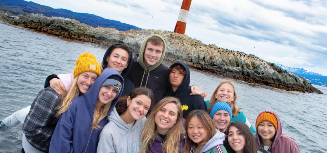 A group os students smile on a boat in front of Les Eclaireurs Lighthouse.