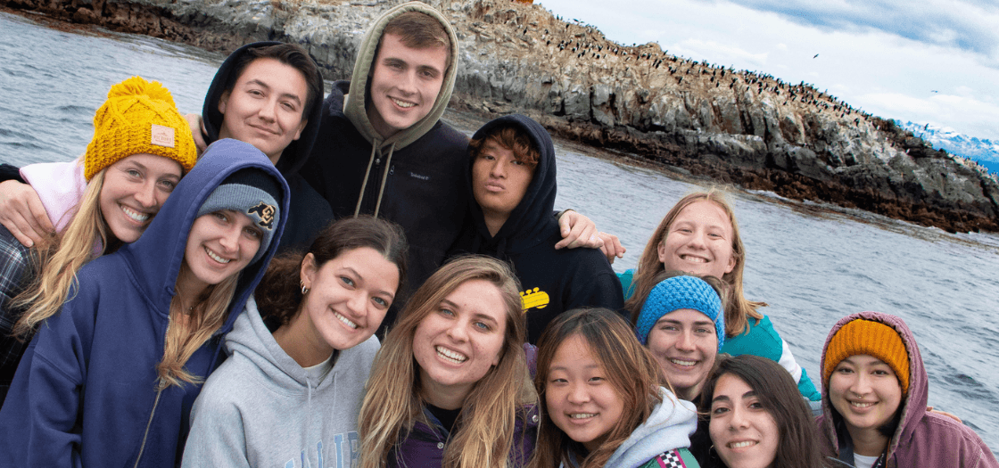 A group os students smile on a boat in front of Les Eclaireurs Lighthouse.