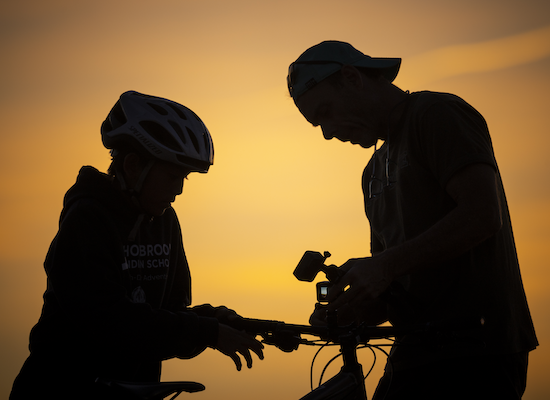 A silhouette of a student and a filmmaker looking into a camera against the backdrop of a sunset